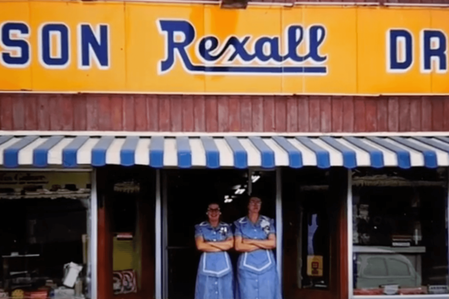 Two waitresses in blue uniforms under the striped awning and Rexall sign